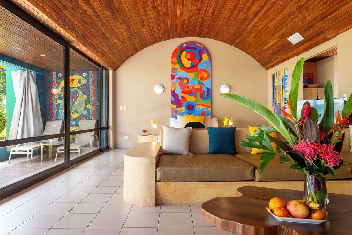 A modern living room with a curved wooden ceiling, a colorful abstract painting, a sofa with cushions, a coffee table with fruit, and a large window with a view of the outdoors.