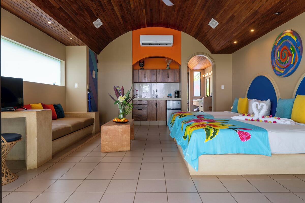 A modern hotel room with a wooden ceiling, a kitchen in the background, a bed with a colorful bedspread, and a TV on a wooden table.