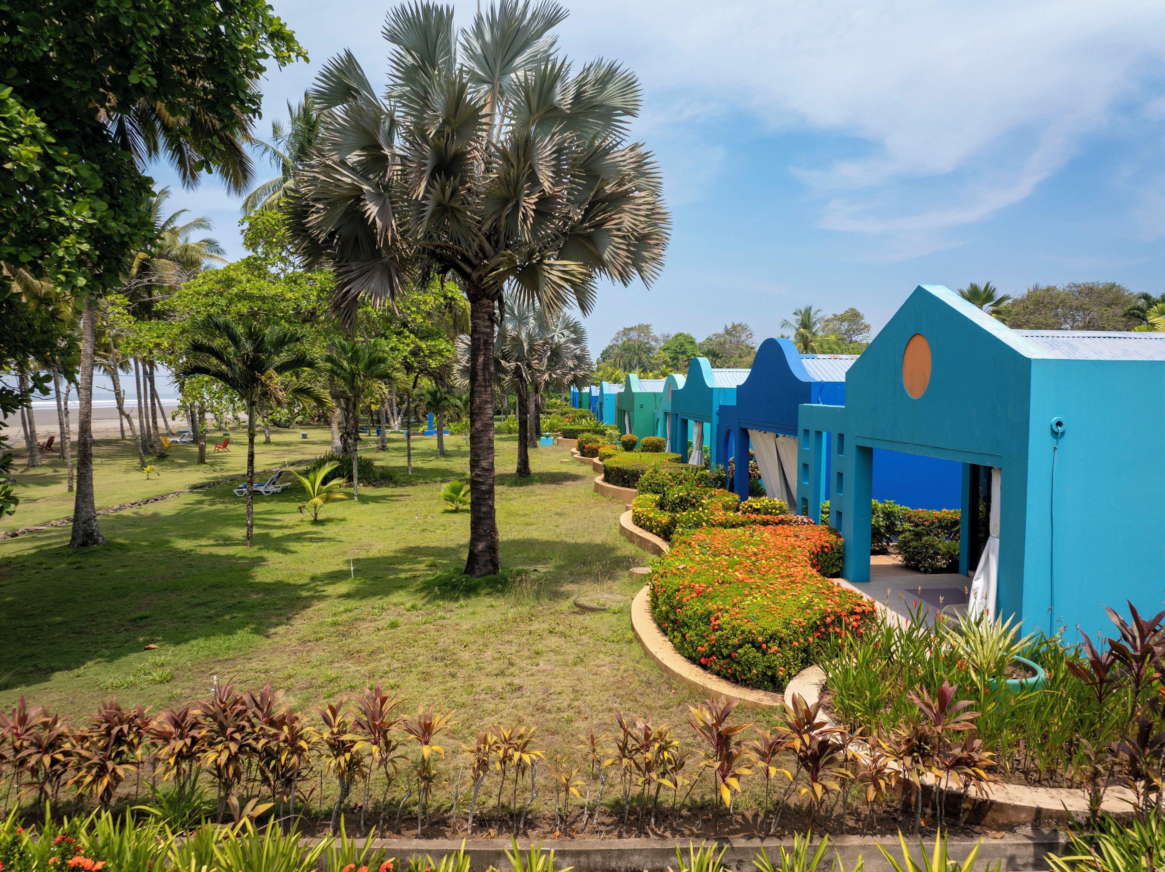A row of blue-roofed buildings with a well-maintained garden and palm trees in front.