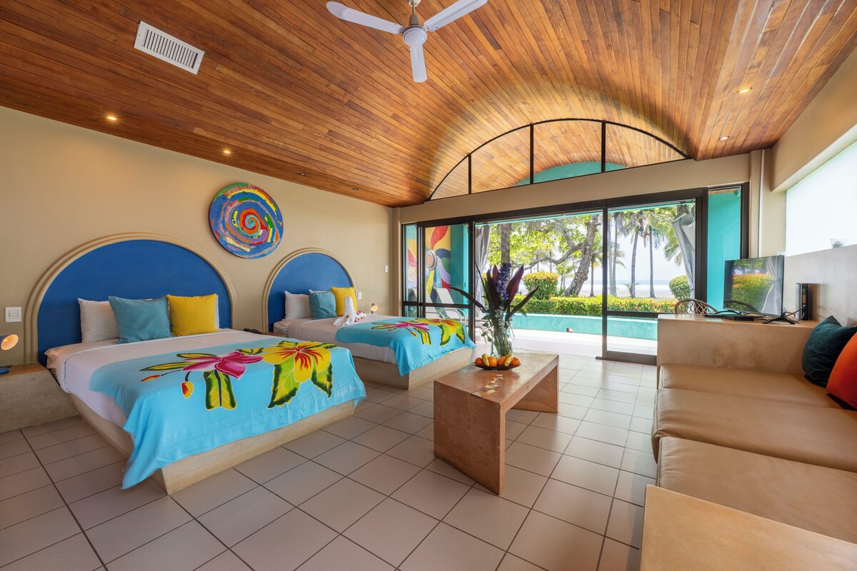 A hotel room with two beds, a sofa, a ceiling fan, and a large window with a view of palm trees.