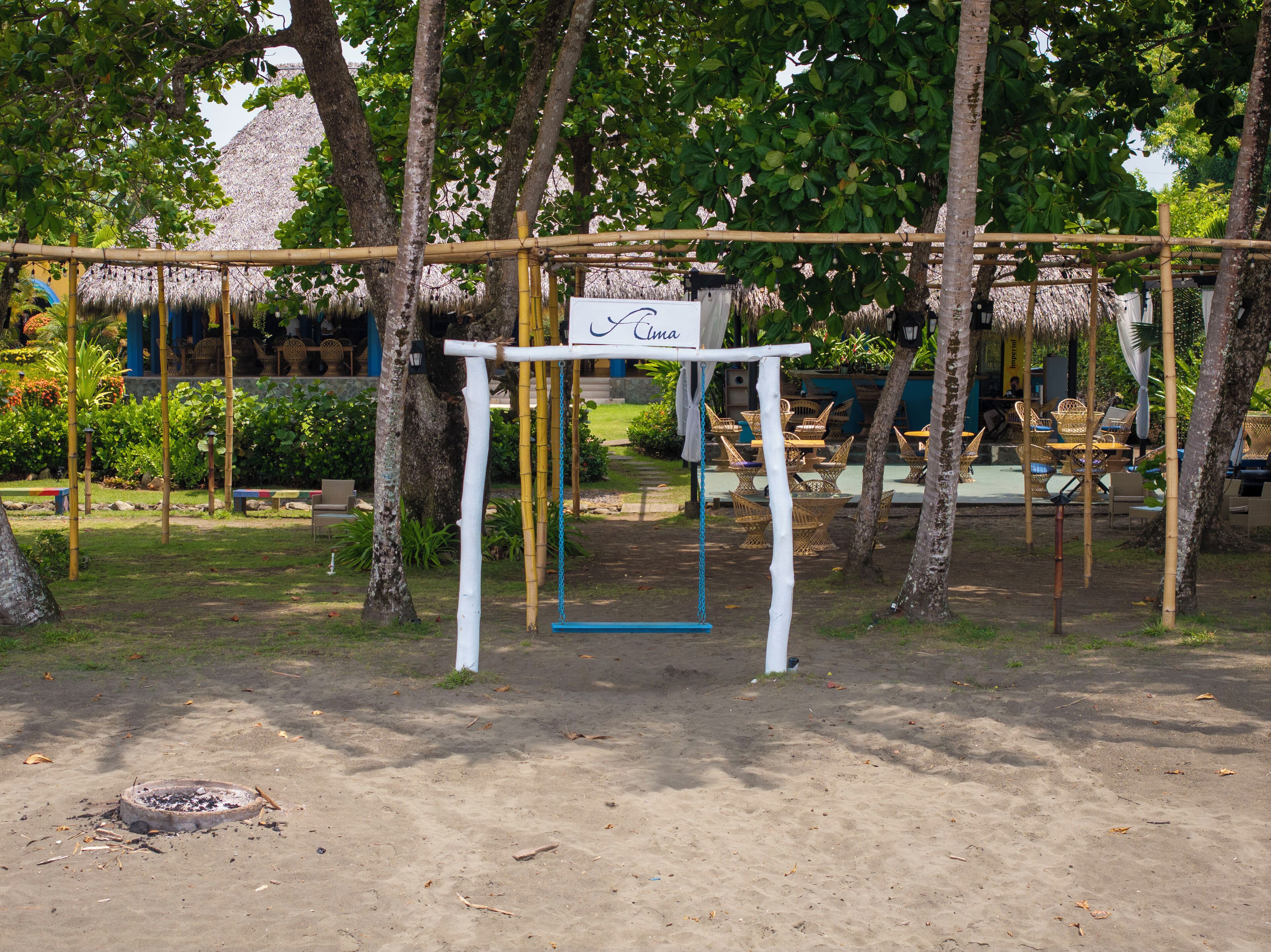 A beachside area with a swing set, a sign reading "Cima," and thatched-roof structures in the background.