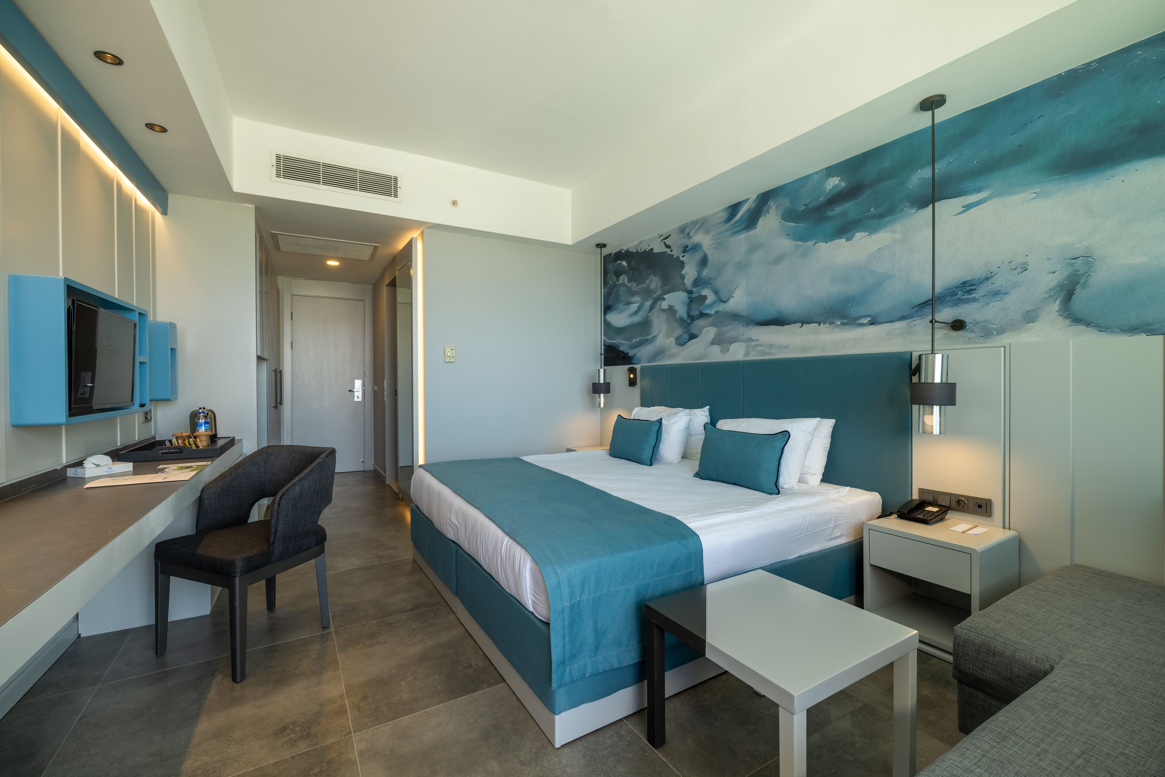 A modern hotel room with a large bed, a desk with a chair, a small table, and a nightstand with a lamp.