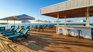 Terraza de madera con tumbonas y sombrillas, una zona de bar y vistas al mar.