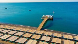 Vista aérea de un resort frente a la playa con un muelle que se adentra en el mar, mostrando tumbonas y sombrillas en zonas sombreadas.