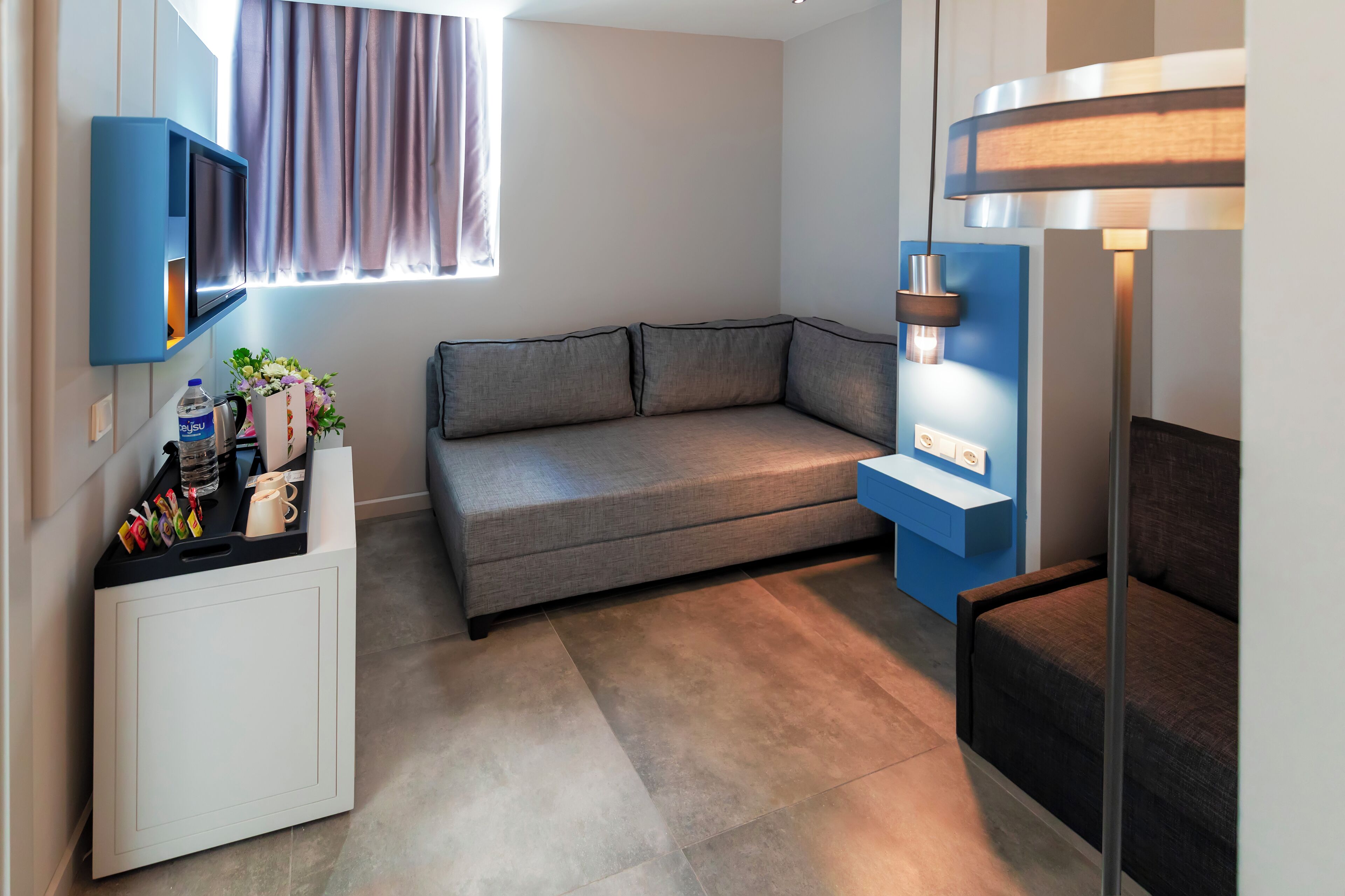 Una sala de estar moderna con un sofá gris, un mueble blanco pequeño, una mesita de noche azul y una lámpara de pie.