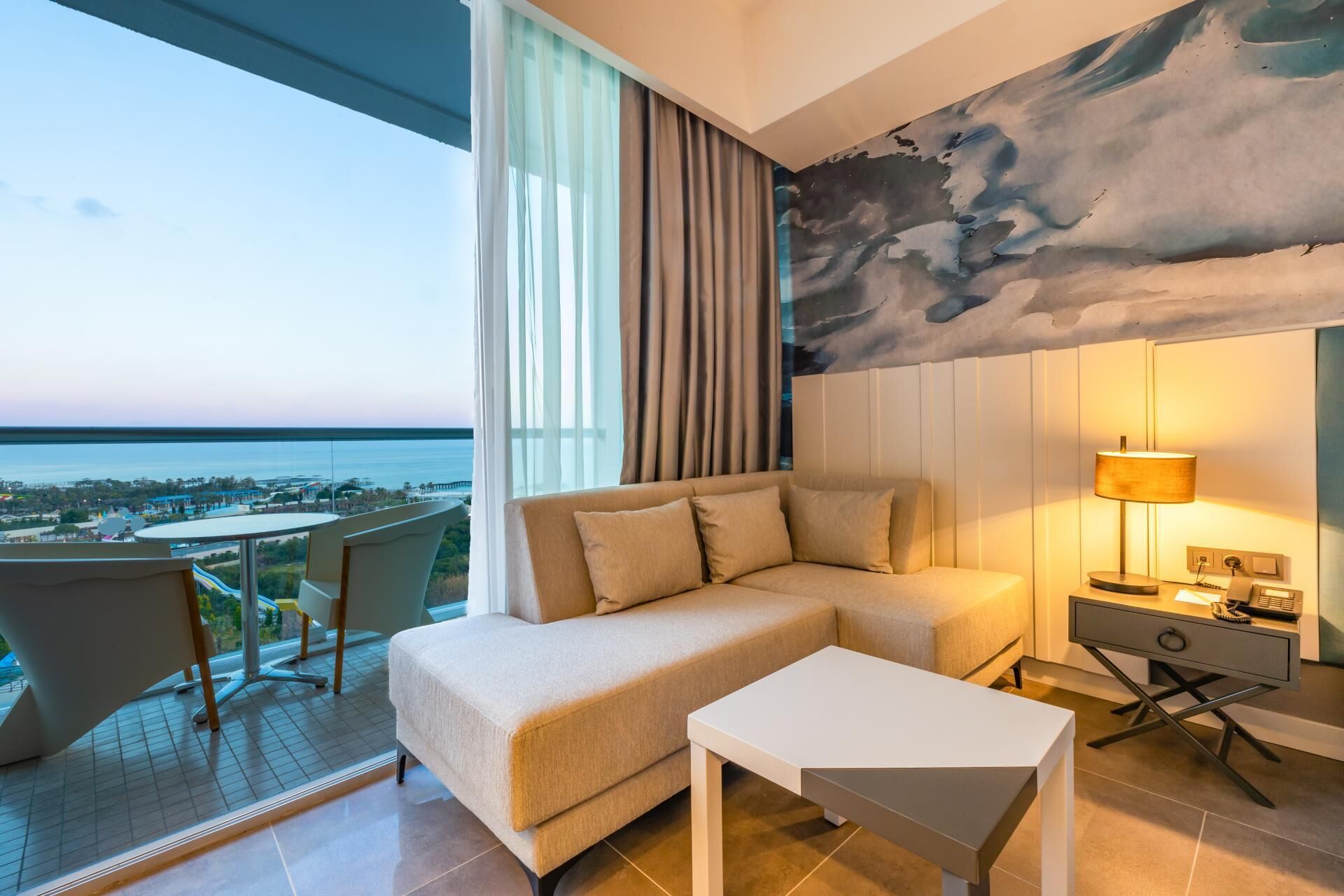 Una habitación de hotel moderna con sofá, una mesita, un escritorio con lámpara y vista al mar.