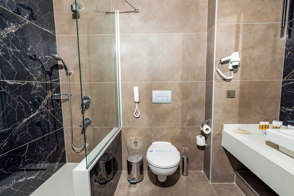 Un baño moderno con ducha envuelta en vidrio, inodoro, cesto de basura y tocador con lavamanos y artículos de aseo.
