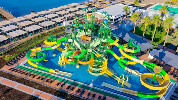 Vista aérea de un parque acuático con múltiples toboganes y piscinas, rodeado de sillones de playa y palmeras.