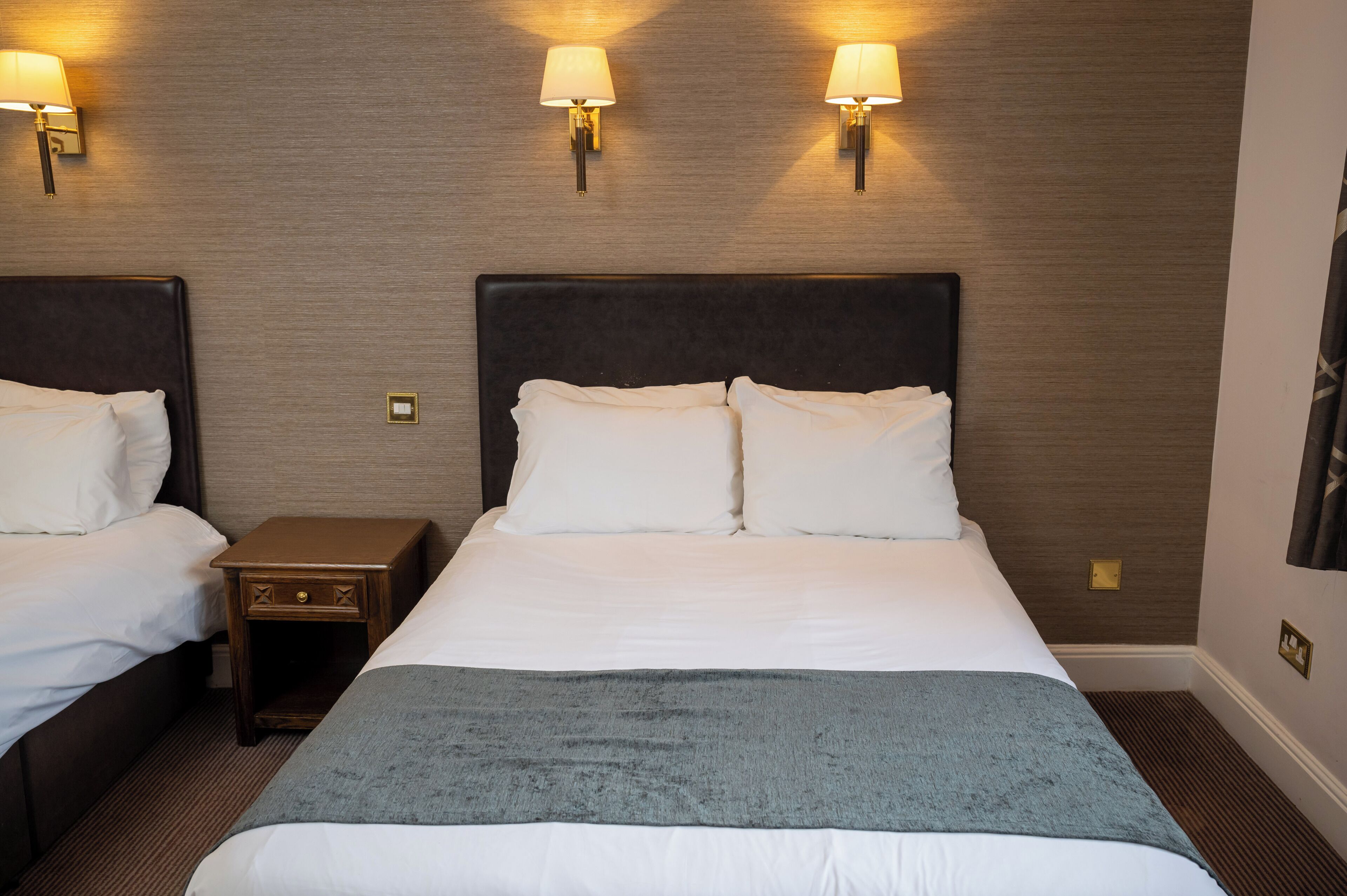 A hotel room with two beds, each with white linens and a dark headboard. There are wall-mounted lamps on either side of the beds, and a small wooden nightstand between them.