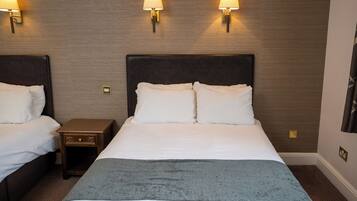 A hotel room with two beds, each with white linens and a dark headboard. There are wall-mounted lamps on either side of the beds, and a small wooden nightstand between them.
