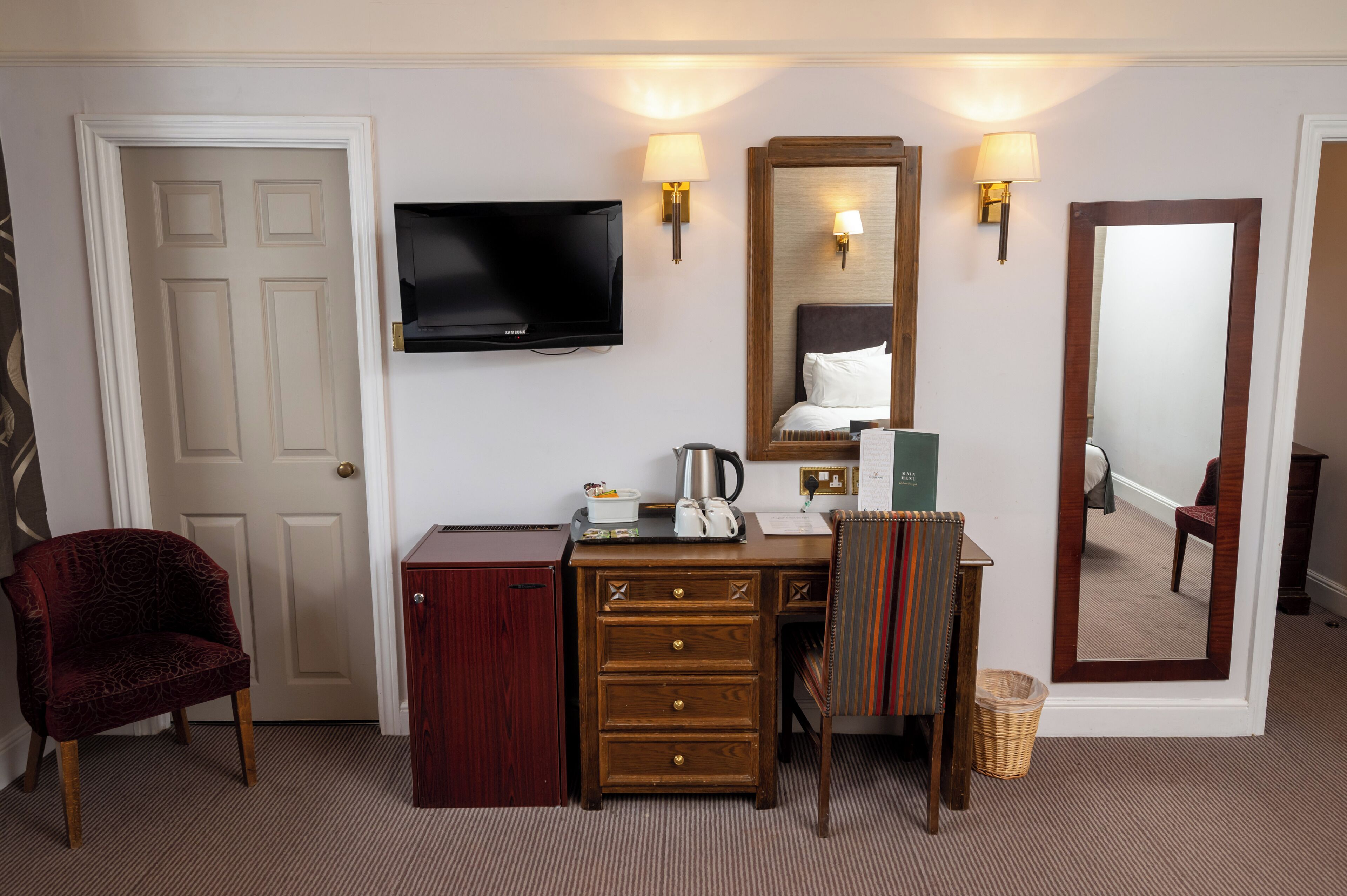 Habitación de hotel con televisor, escritorio con cajones, una silla, espejo y lámpara.