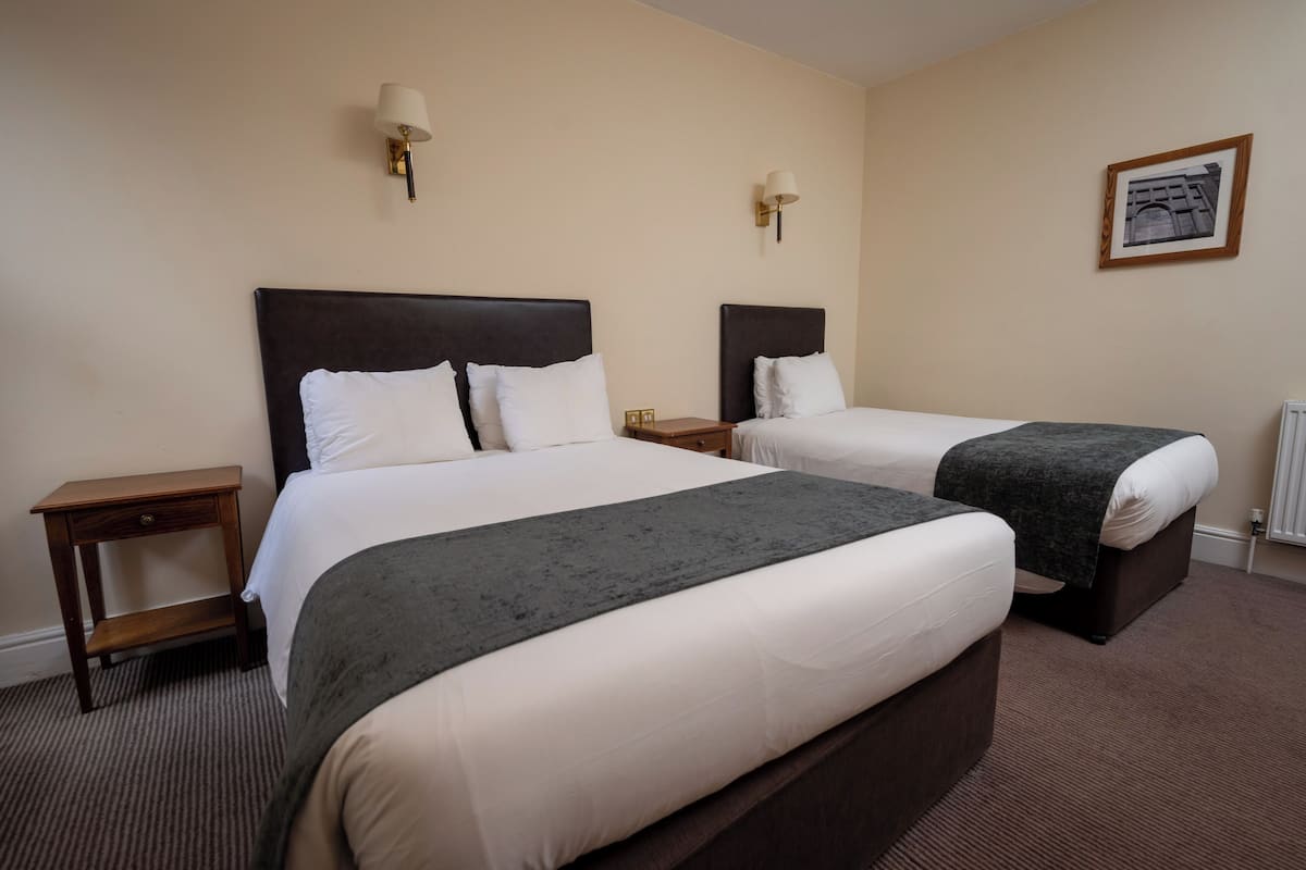 A hotel room with two beds, each with a dark headboard and white bedding. There are two bedside tables with lamps, a framed picture on the wall, and a carpeted floor.