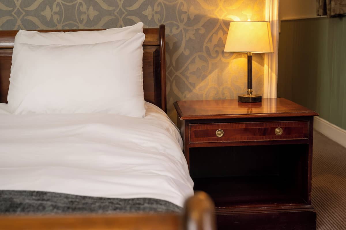 A neatly made bed with white linens, a wooden nightstand with a lamp, and a patterned wallpaper background.
