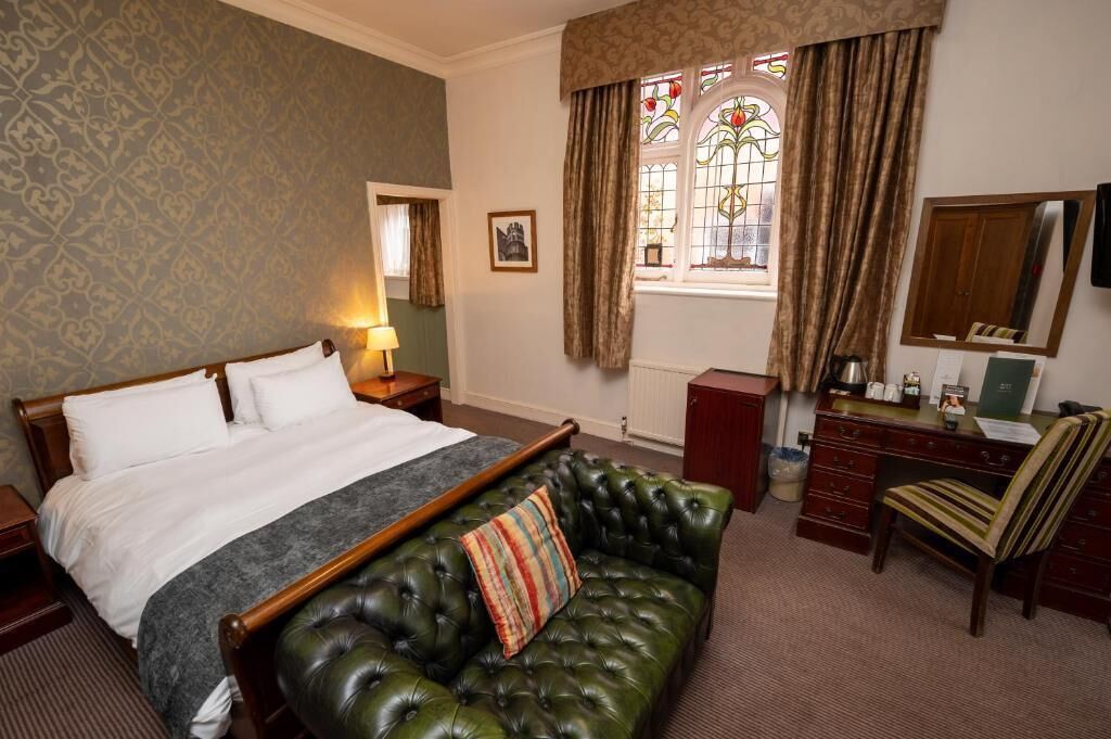 A hotel room with a large bed, a green leather sofa, a desk with a chair, a dresser, and a window with curtains.
