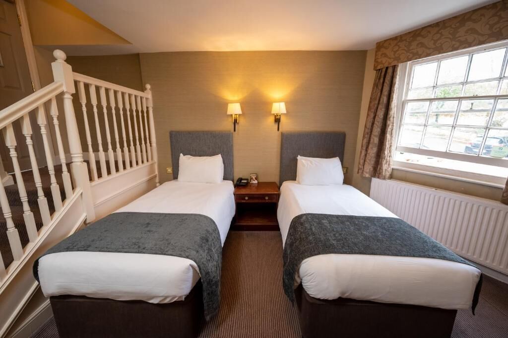 A hotel room with two beds, a wooden staircase, a window with curtains, and two wall-mounted lamps.