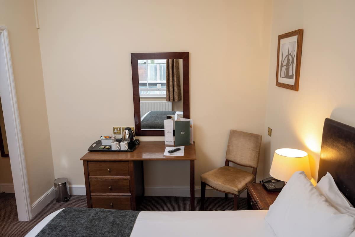 A hotel room with a bed, a desk with a chair, a lamp, a framed picture, and a window with curtains.