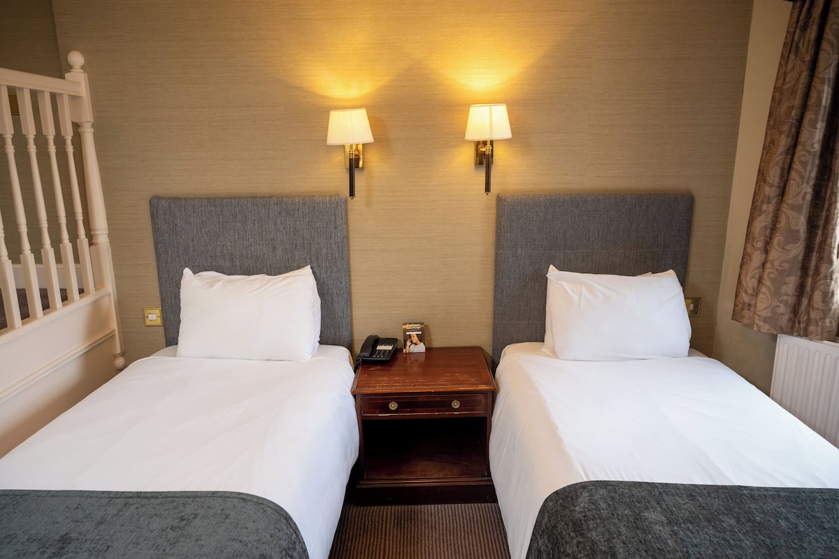 A hotel room with two single beds, a nightstand with a phone and a small table, and two wall-mounted lamps.