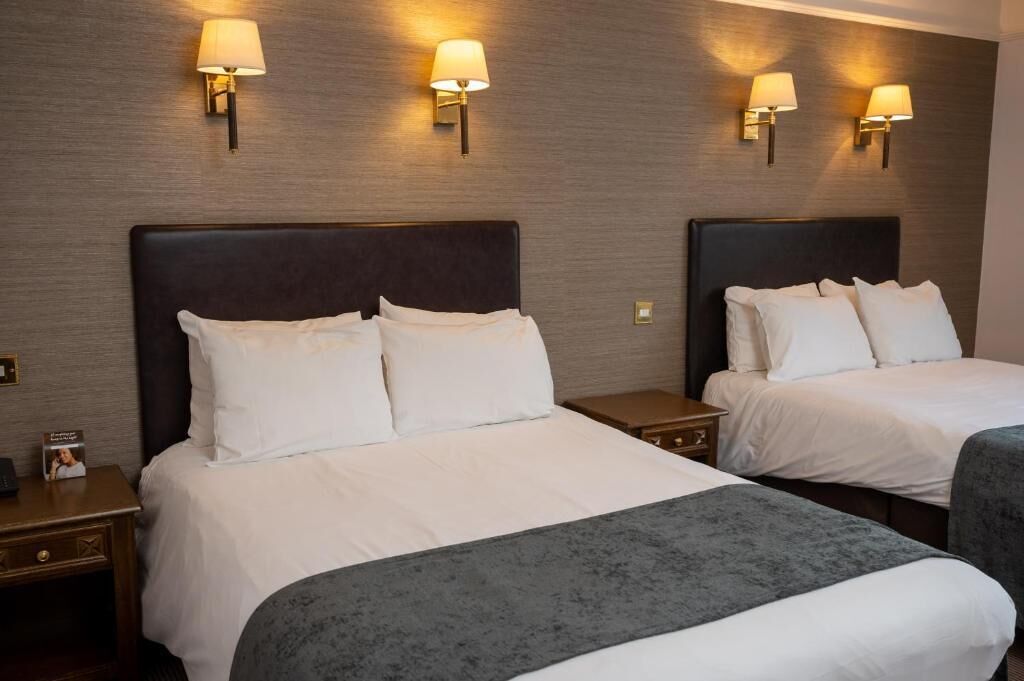 Habitación de hotel con dos camas, cada una con ropa blanca y cabeceros oscuros. Hay lámparas de pared a cada lado de las camas.