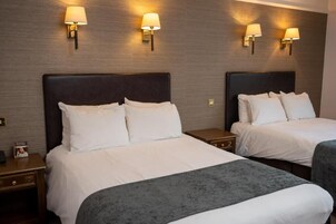 Quarto de hotel com duas camas, cada uma com roupa de cama branca e cabeceira escura. Há luminárias fixadas na parede, uma de cada lado das camas.