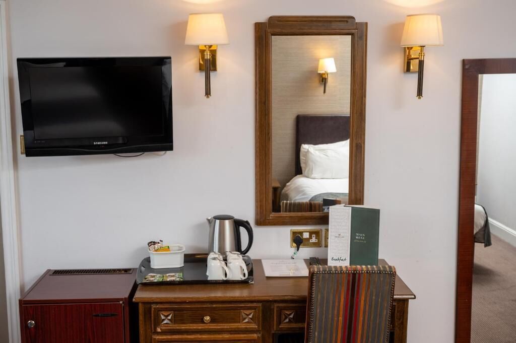 A hotel room with a flat-screen TV, a wooden desk with a kettle, a tray with cups, a mirror, and two wall-mounted lamps.