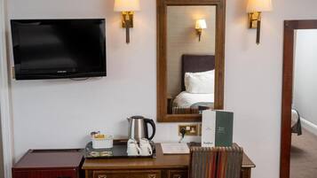 A hotel room with a flat-screen TV, a wooden desk with a kettle, a tray with cups, a mirror, and two wall-mounted lamps.