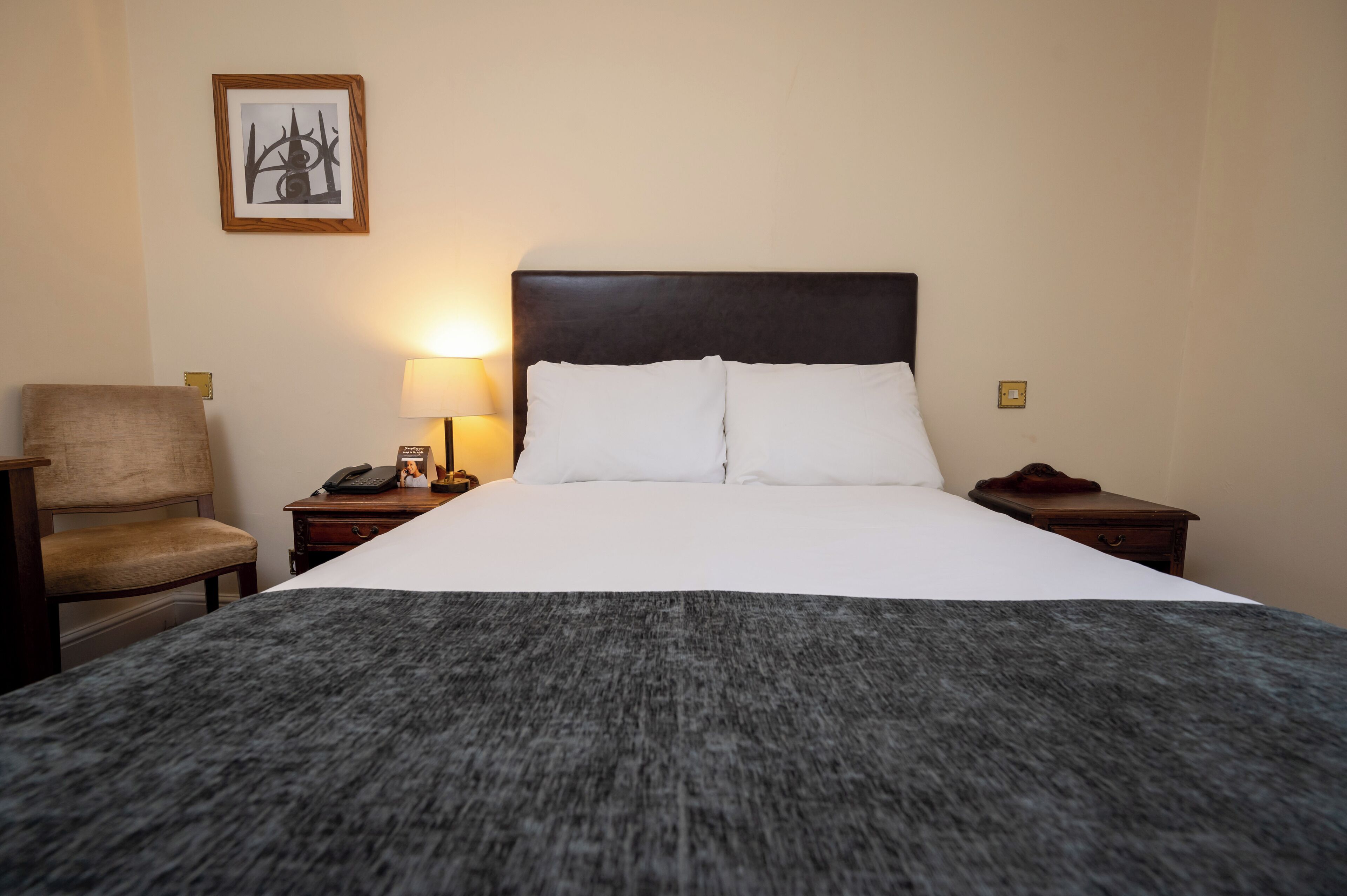A hotel room with a large bed, a chair, a nightstand with a lamp, and a framed picture on the wall.