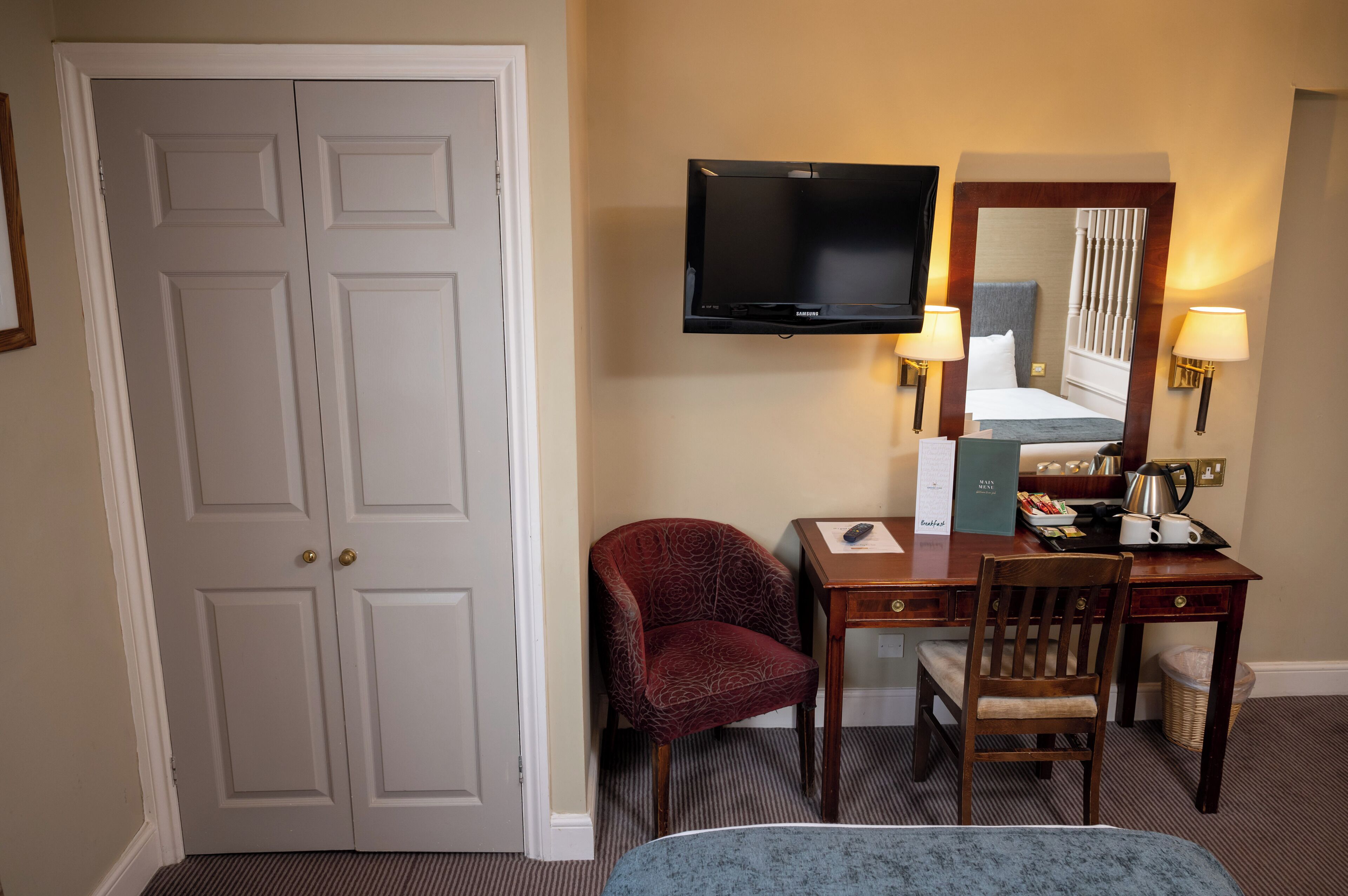 Een hotelkamer met een grote witte deur, een flatscreen televisie aan de muur, een bureau met een stoel, een bed met hoofdeinde en twee wandlampen.