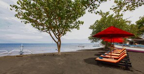 Une plage avec des chaises longues et un parasol rouge, un arbre et des bateaux sur l’eau.