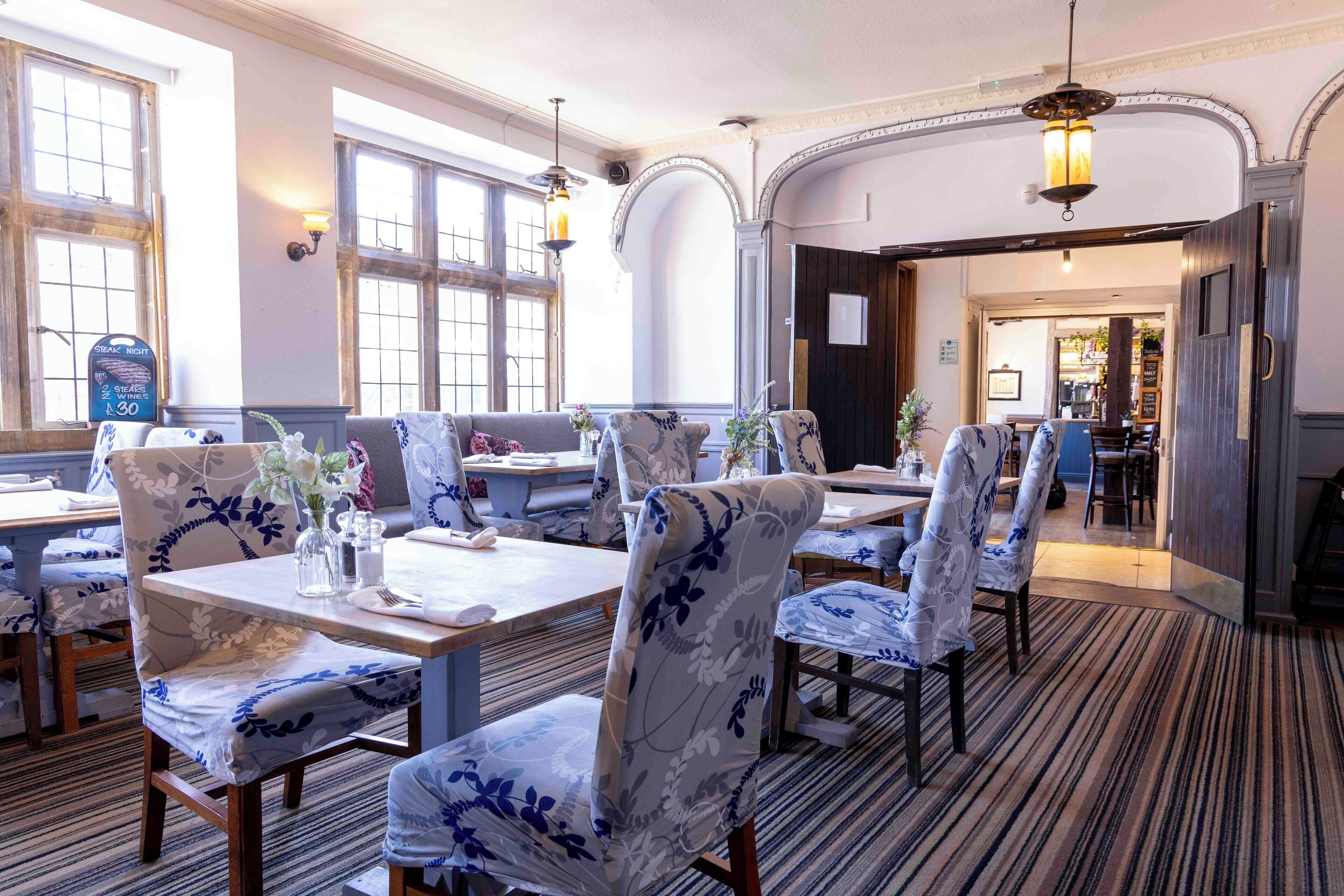 A dining area with tables and chairs, large windows, and a carpeted floor.