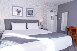 Een net opgemaakt bed met witte lakens en een grijze plaid, een houten stoel en een nachtkastje met een lamp.