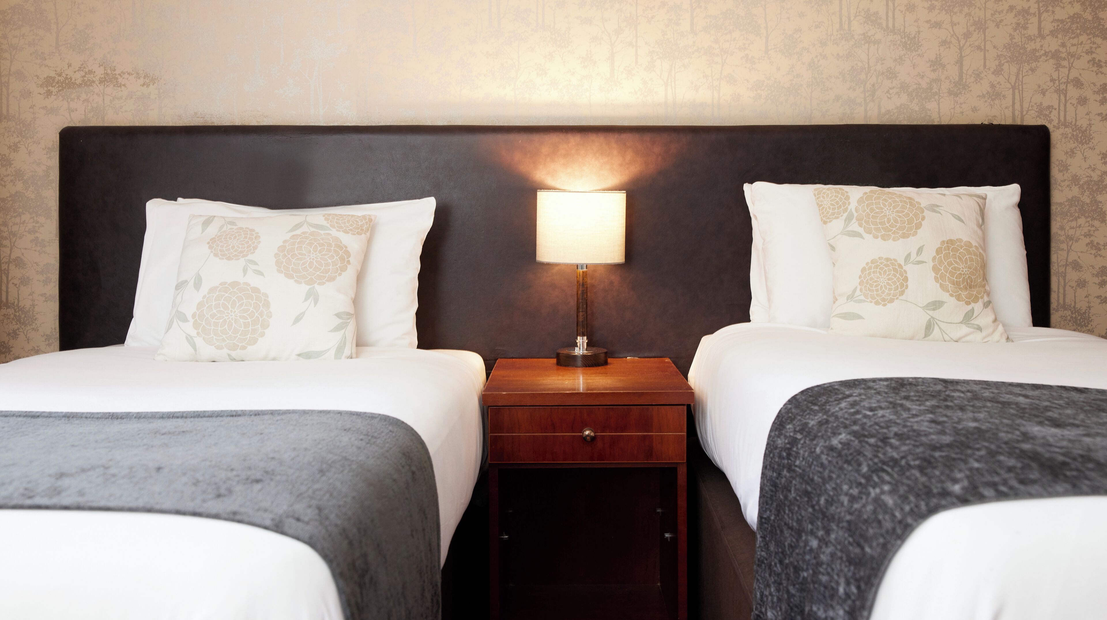 A hotel room with two beds, each with floral-patterned pillows, a nightstand with a lamp, and a small wooden drawer.