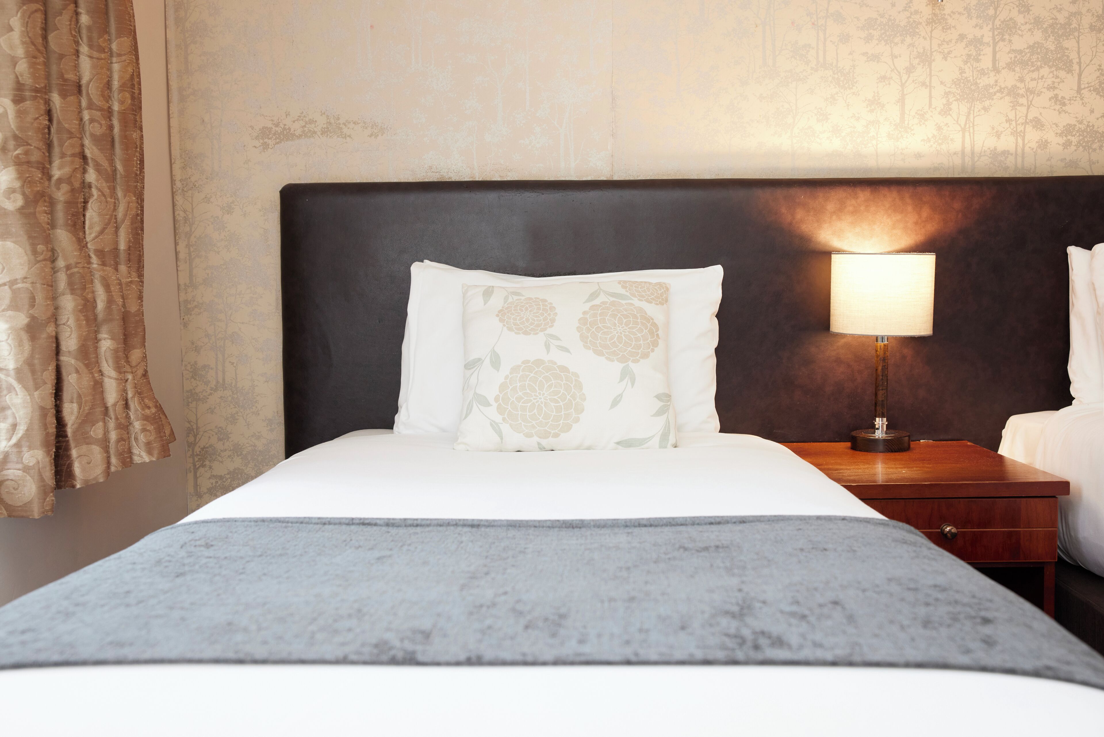 A neatly made bed with a grey blanket, a dark headboard, and a bedside table with a lamp.