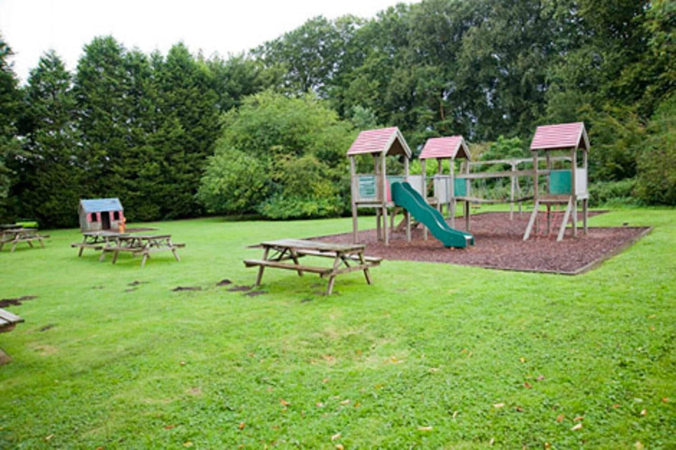 Une aire de jeux avec un toboggan et des structures d’escalade, entourée de tables de pique-nique et d’une petite structure ressemblant à une maison.