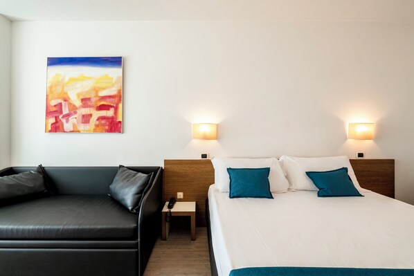 A hotel room with a bed, a black leather sofa, a nightstand with a phone, and a painting on the wall. - Hotel Cristallo (Rimini)