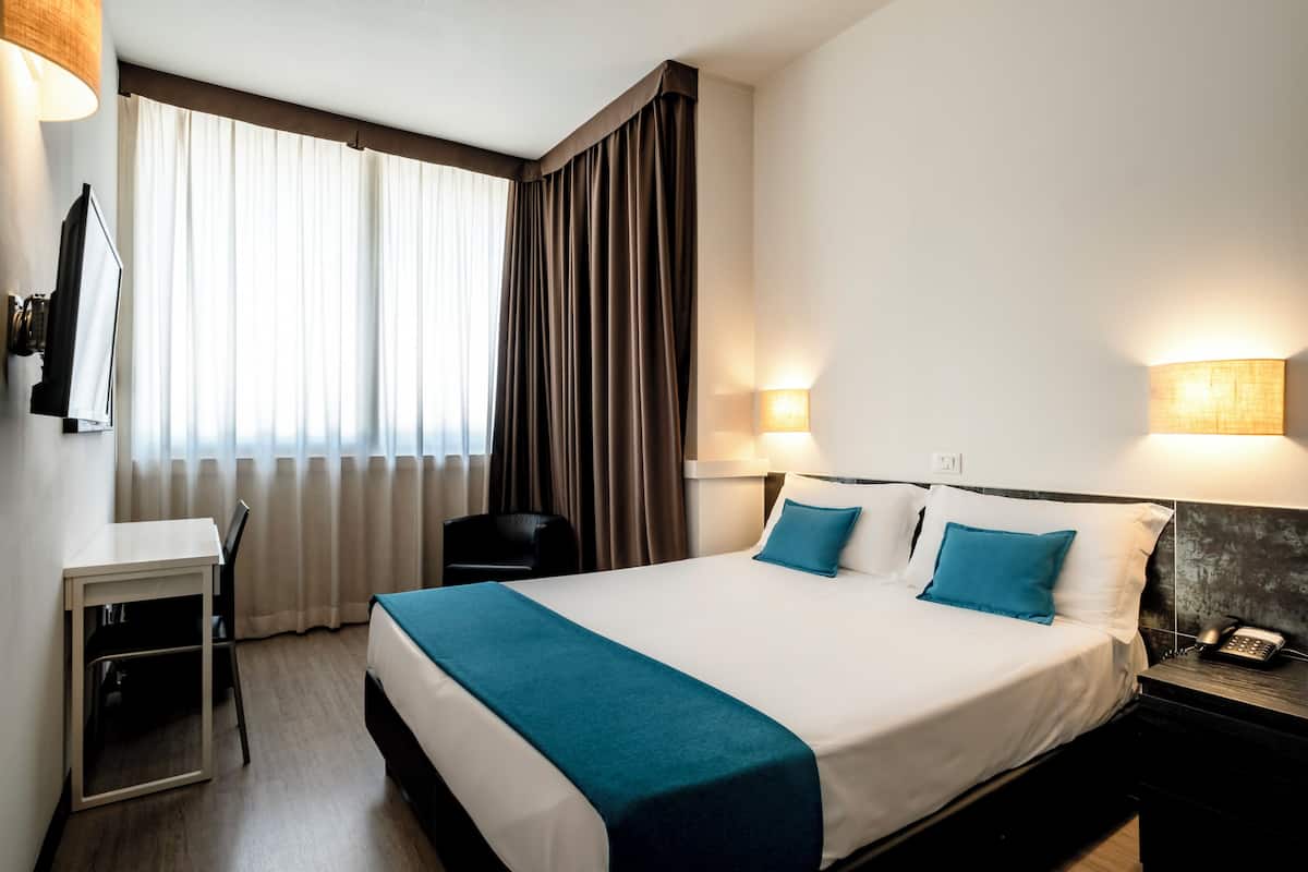 A hotel room with a bed, bedside table, lamp, and a desk with a chair.