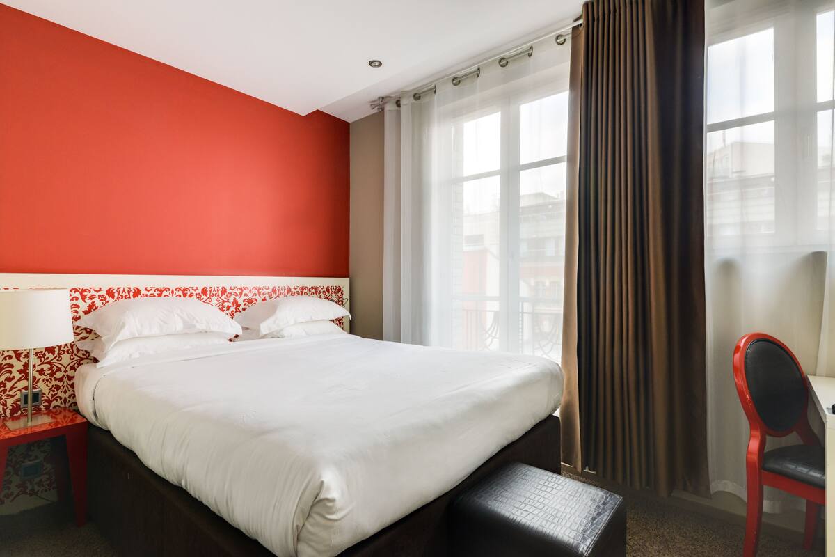 Une chambre d’hôtel avec un grand lit, un mur d’accent rouge, une fenêtre avec des rideaux et un fauteuil rouge.