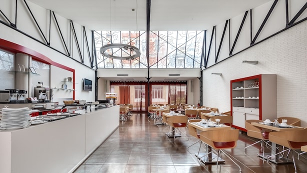 Un comedor moderno con mesas y sillas largas, una barra de cocina con vajilla y un amplio ventanal con diseño geométrico.