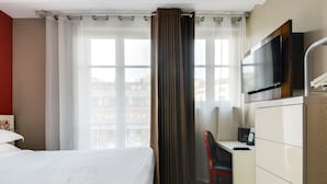 Habitación de hotel con cama, un escritorio y silla, televisión y vistas a edificios a través de amplias ventanas.