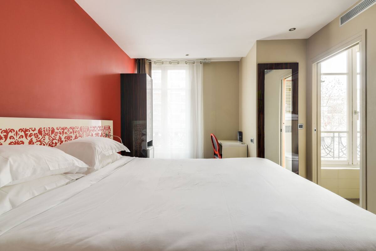 Une chambre d’hôtel avec un grand lit, un bureau et une chaise. Elle est dotée d’une fenêtre avec des rideaux, d’une porte donnant sur un balcon et d’un mur d’accent de couleur rouge.