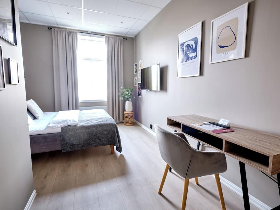 A modern hotel room with a bed, desk, chair, TV, and artwork on the wall.
