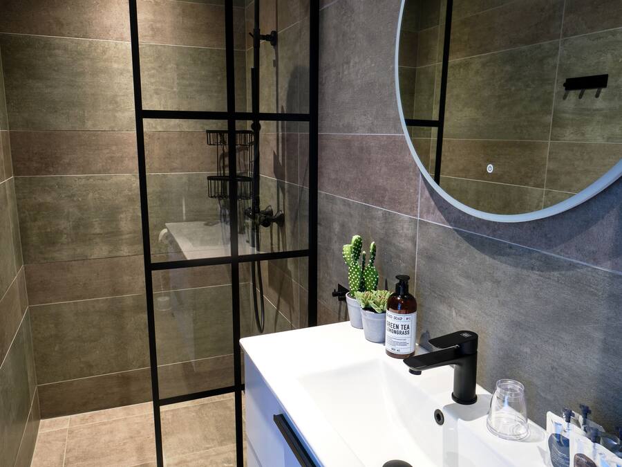 Een moderne badkamer met een glazen douchecabine, een witte wastafel met een zwarte kraan en een ronde spiegel.