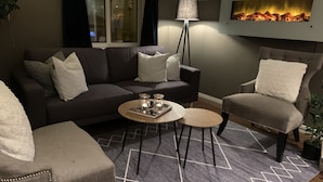 A modern living room with a dark-colored sofa, two armchairs, a coffee table, and a floor lamp. A fireplace is visible on the wall, and a window with curtains is present. - Moss Hotel & Apartments (Moss)
