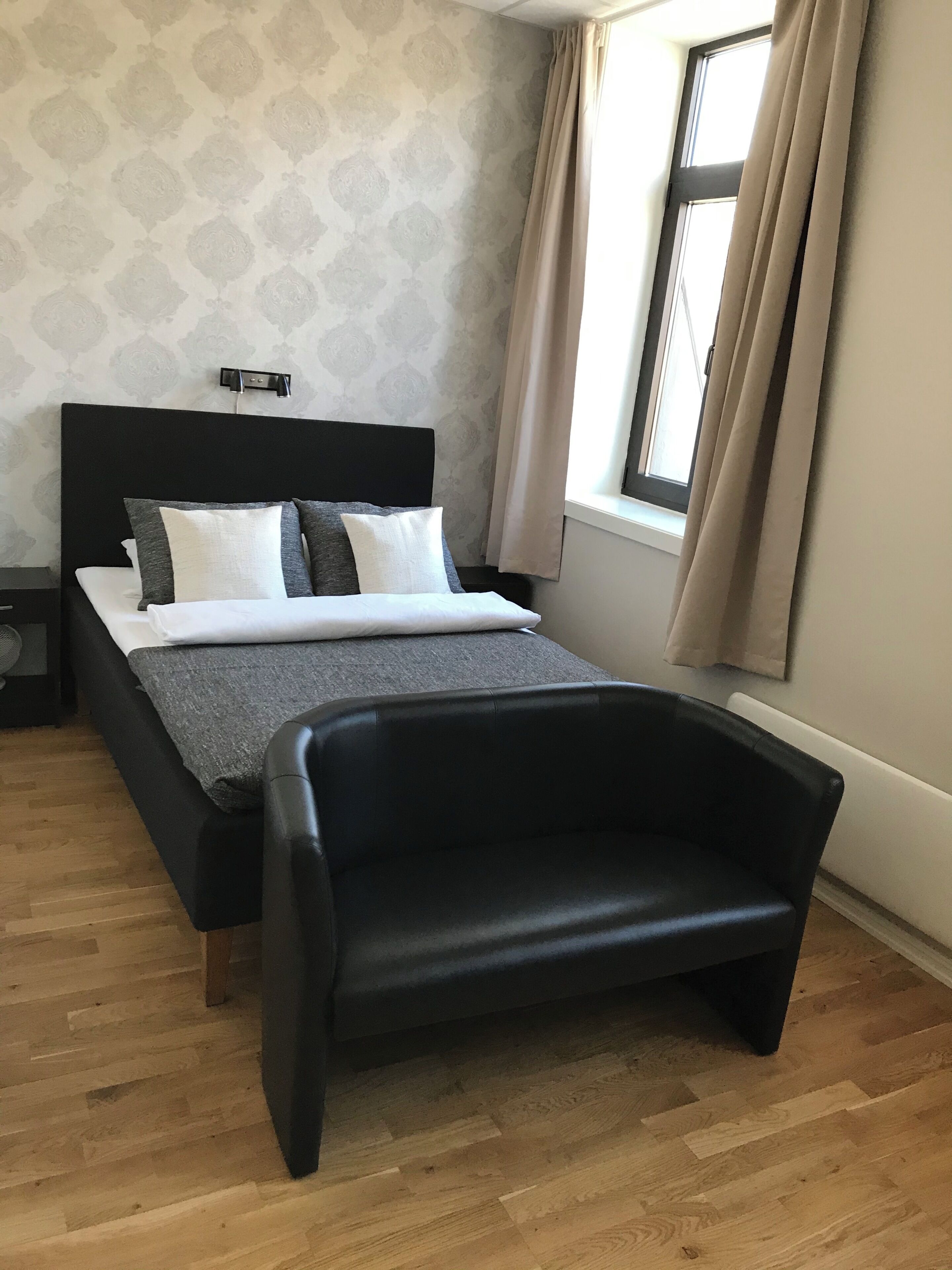 Une chambre à coucher moderne avec un lit, un fauteuil noir, une table de chevet et une fenêtre avec des rideaux.