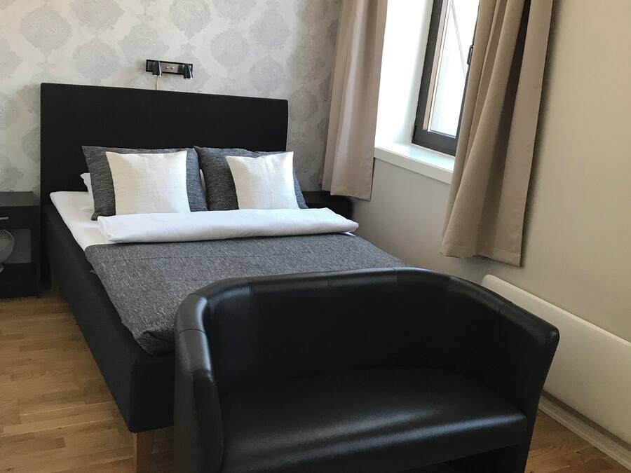 Une chambre à coucher moderne avec un lit, un fauteuil noir, une table de chevet et une fenêtre avec des rideaux.