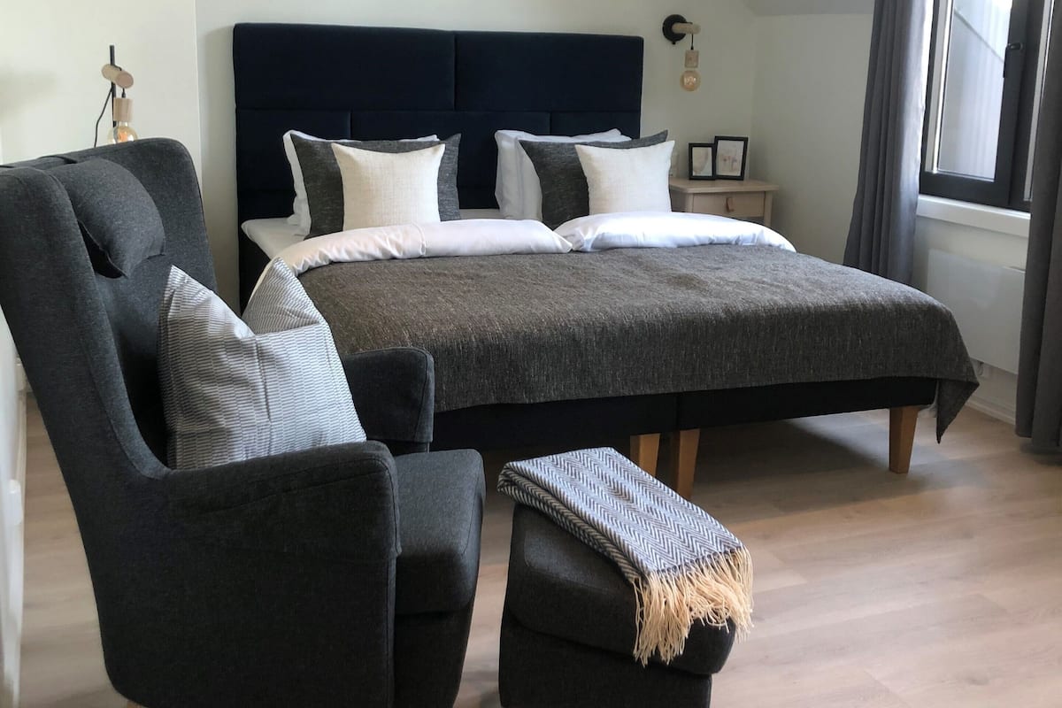Un dormitorio moderno con una cama grande, un sillón tapizado oscuro, un pequeño otoman y piso de madera.
