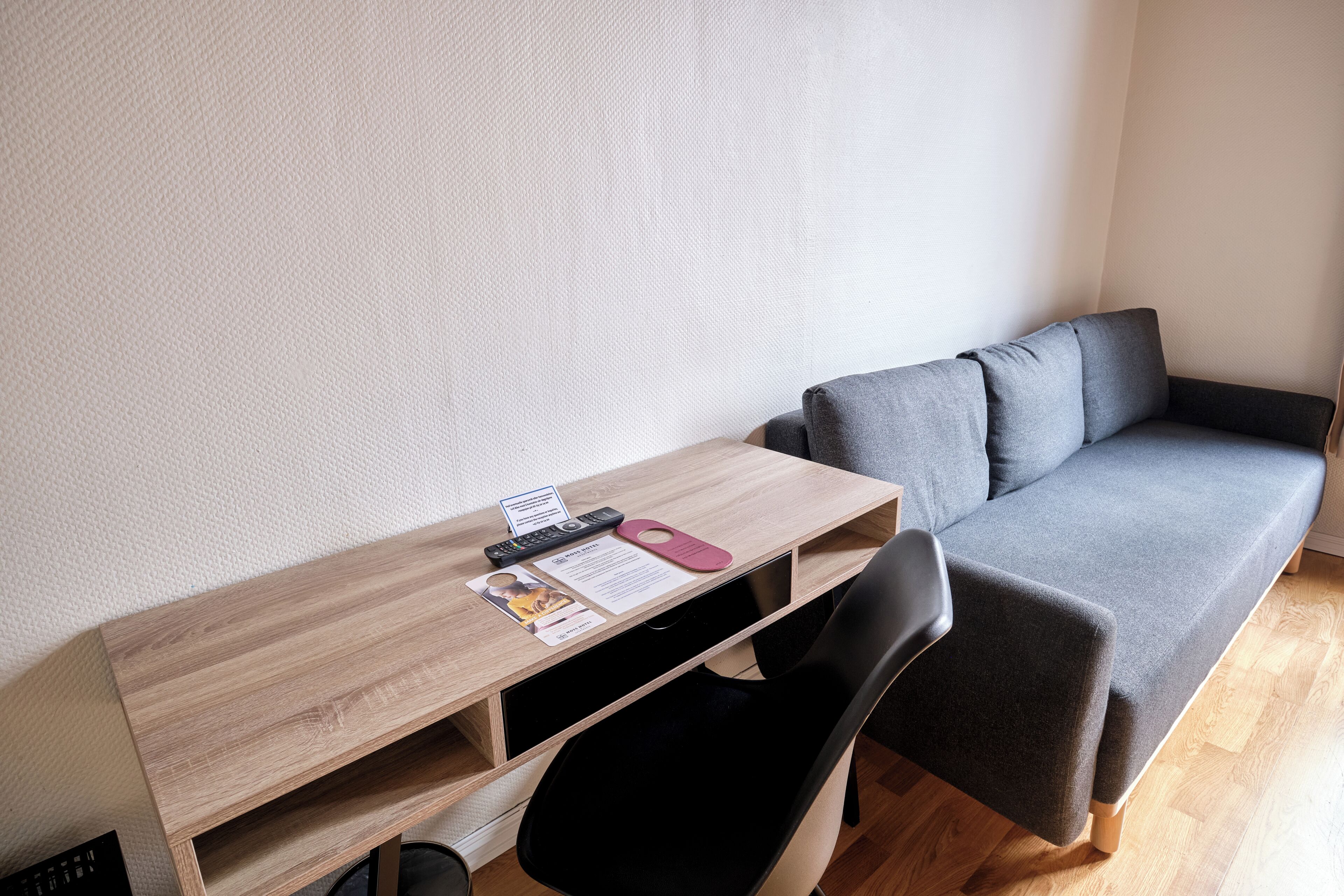 Una habitación pequeña con un escritorio de madera, un sofá gris, una silla negra y un control remoto.