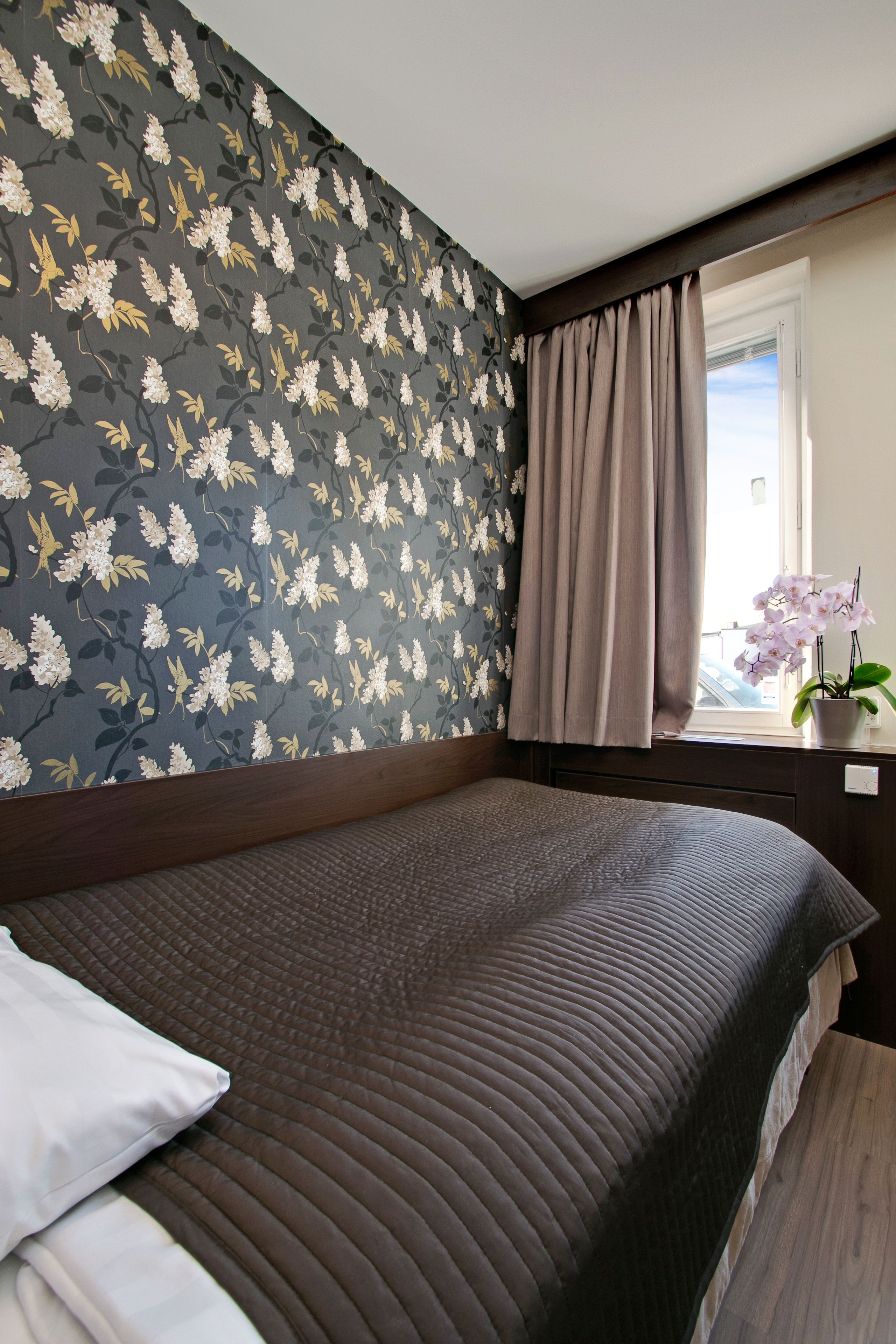 A bedroom with a floral wallpaper, a bed with a dark headboard, a window with a view, and a potted orchid on the windowsill.
