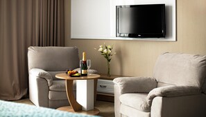 A hotel room with two armchairs, a small round table with a bottle of champagne, a glass, and a fruit platter. A flat-screen TV is mounted on the wall, and a vase with white flowers is on a side table. - Flowers Hotel (Chisinau)