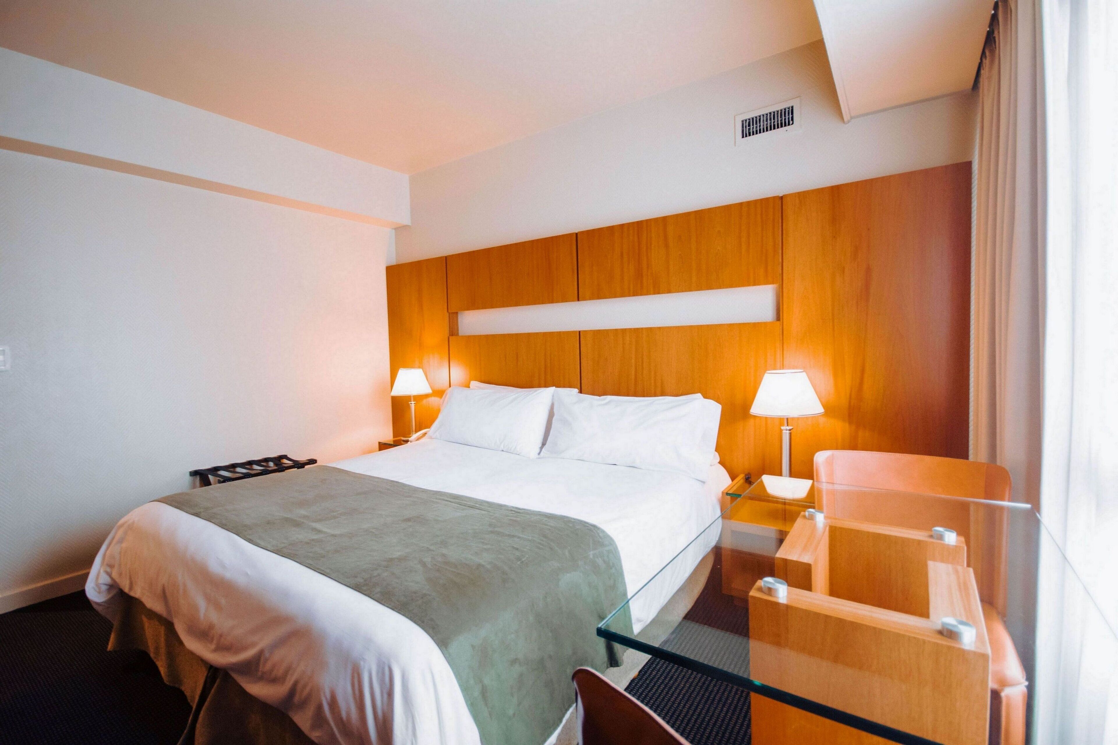 A hotel room with a bed, bedside lamps, a glass table, and wooden paneling on the wall.
