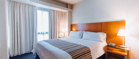 A hotel room with a large bed, a nightstand with a lamp, and a view of the outdoors through the window.